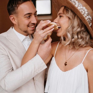 Puede incluir: Una pareja compartiendo una galleta, la mujer lleva un sombrero marr&oacute;n de ala ancha con una banda blanca y un vestido blanco, el hombre lleva un traje beige claro.