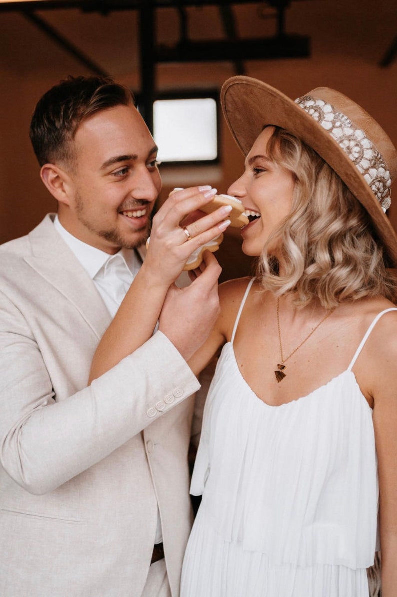 Puede incluir: Una pareja compartiendo una galleta, la mujer lleva un sombrero marr&oacute;n de ala ancha con una banda blanca y un vestido blanco, el hombre lleva un traje beige claro.