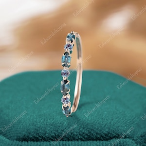 Puede incluir: Un delicado anillo de banda de oro con una fila de piedras preciosas azules y moradas. El anillo está colocado sobre un fondo de terciopelo turquesa.