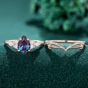 May include: A rose gold ring set featuring a pear-cut color-changing center stone flanked by small marquise-cut white stones, paired with a matching V-shaped chevron band that has delicate cut-out details and small accent stones on the sides.