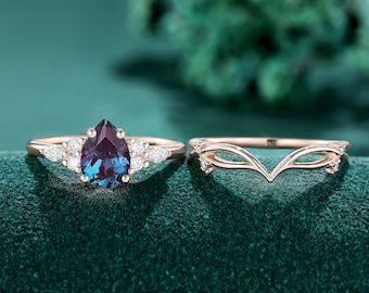 May include: A rose gold ring set featuring a pear-shaped color-changing center stone flanked by small marquise and round white stones, accompanied by a matching V-shaped chevron band with delicate cut-out details and small accent stones.