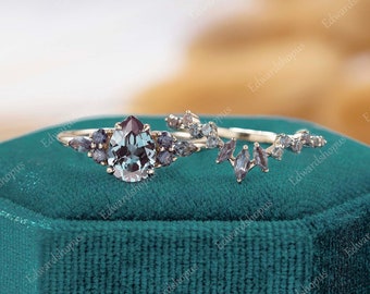 May include: A pear-shaped gemstone ring with a delicate band and a matching band with a row of smaller gemstones. The ring is set in gold and features a blue-green gemstone with a hint of purple.