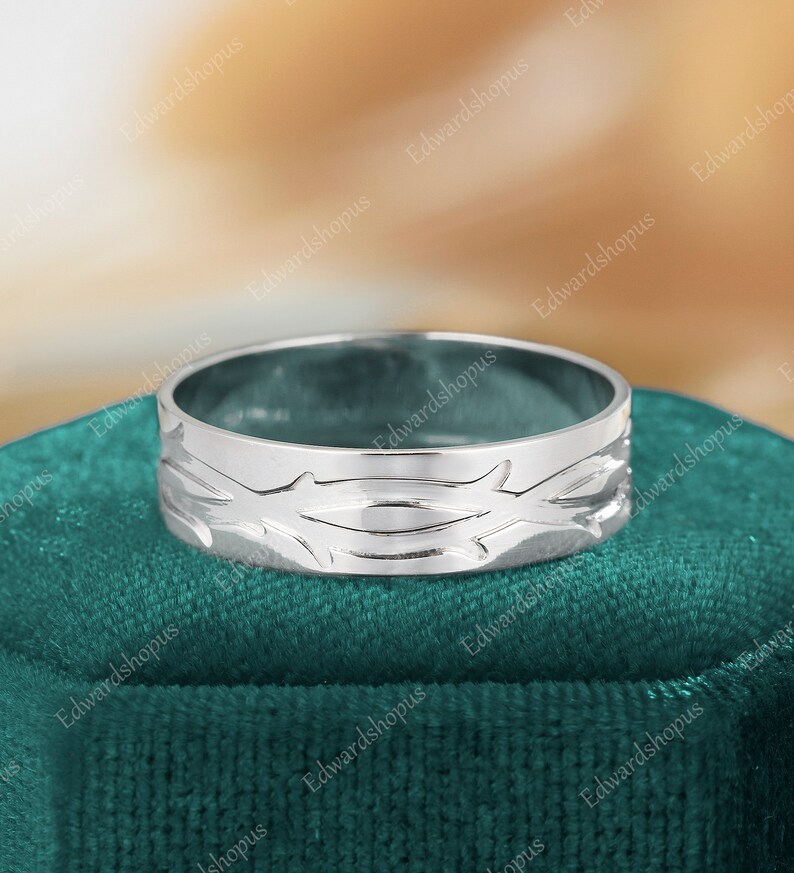 K&ouml;nnte beinhalten: Ein silberner Bandring mit einem detaillierten Design aus ineinander verschlungenen &Auml;sten. Der Ring wird auf einem gr&uuml;nen Samtstoff pr&auml;sentiert.