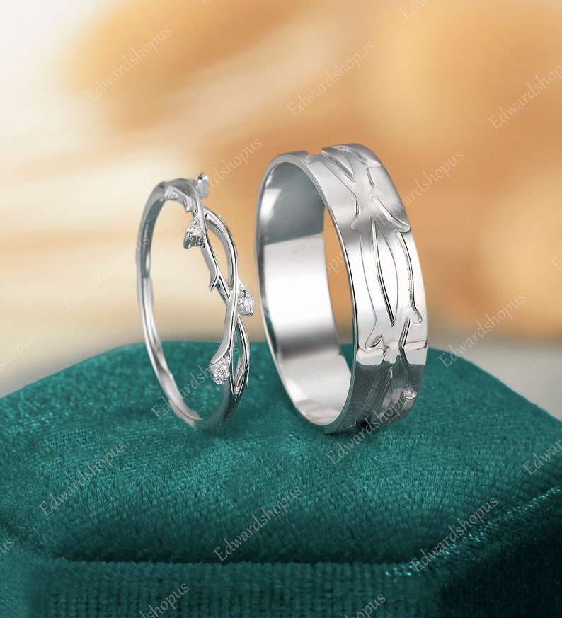 K&ouml;nnte beinhalten: Zwei silberne Eheringe auf einem gr&uuml;nen Samtkissen. Ein Ring zeigt ein filigranes Design aus ineinander verschlungenen &Auml;sten mit kleinen, klaren Steinen. Der andere Ring hat ein strukturiertes Design aus ineinander verschlungenen &Auml;sten.