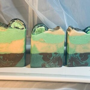 Puede incluir: Tres jabones artesanales con capas de colores verde, beige y marrón. La parte superior de cada jabón tiene un diseño oscuro y texturizado. Los jabones se exhiben en una bandeja blanca.