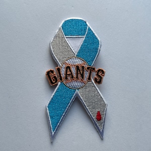 Puede incluir: Una cinta azul y blanca con un logotipo de béisbol y la palabra "GIANTS" en negro. Una sola gota de sangre roja está en la parte inferior de la cinta.