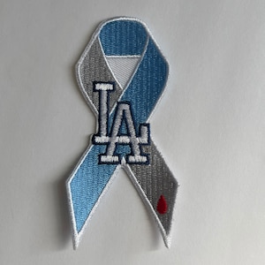 May include: Embroidered ribbon patch in light blue and gray, featuring the letters "LA" in white and blue. A red drop of blood is at the bottom. The patch is likely for awareness or support.