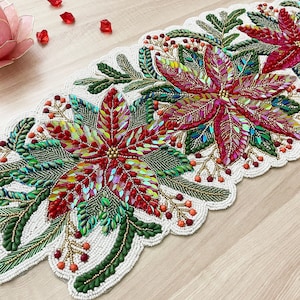 Może przedstawiać: Bielony bieżnik stołowy z koralików z wzorem czerwonych gwiazd betlejemskich z zielonymi liśćmi. Bieżnik jest ozdobiony tęczowymi koralikami w różnych kolorach.