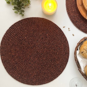 May include: A dining table setting viewed from above. A large, circular, dark brown beaded placemat dominates the scene. Other items include a wooden plate, a bread basket, and a lit candle, creating a warm, inviting atmosphere.