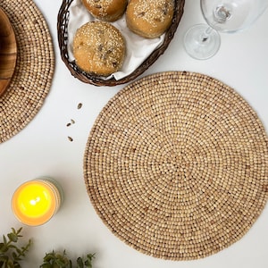 May include: Two round woven placemats made of natural materials, with a third placemat in the background. The placemats are beige and have a circular pattern. There are four bread rolls on a white plate in the background.