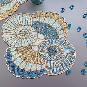 Puede incluir: Un mantel individual de mesa azul, blanco y dorado con un diseño de conchas marinas. El mantel individual está hecho de pequeñas cuentas que se cosen juntas para crear un patrón detallado.