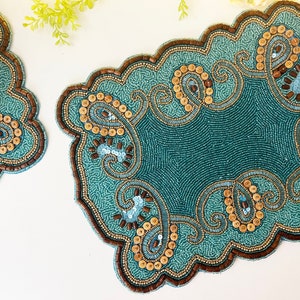 May include: Two teal blue beaded placemats with a paisley design and brown accents. The placemats have a scalloped edge.