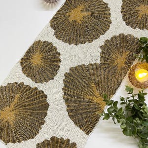 May include: A white beaded table runner with a repeating pattern of gold beaded circles. The circles have a sunburst design.