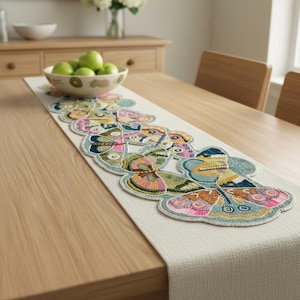 Può includere: Runner da tavolo beige con motivi a farfalla ricamati in vari colori, tra cui rosa, blu, verde e giallo. Il runner è posizionato su un tavolo di legno chiaro, con una ciotola di mele verdi e un vaso di fiori bianchi sullo sfondo.