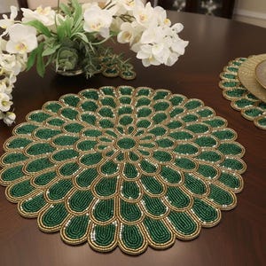 Puede incluir: Un mantel individual redondo con un patrón concéntrico en forma de pétalos, creado con cuentas de color verde oscuro y oro metálico. El diseño irradia desde un punto central, con pequeñas cuentas blancas intercaladas en el intrincado trabajo.