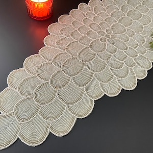 Puede incluir: Un camino de mesa blanco de cuentas con un diseño floral. El camino de mesa está hecho de pequeñas cuentas redondas que se cosen juntas para crear un patrón delicado e intrincado.
