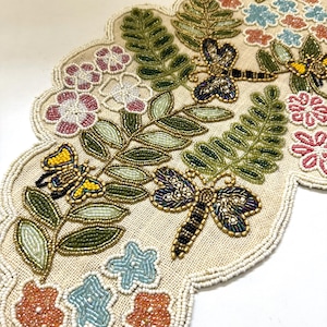 May include: A beige table runner with a beaded design featuring butterflies, dragonflies, and flowers in shades of pink, green, blue, and yellow.