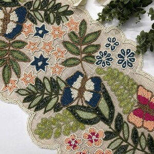 Op de afbeelding: Met kralen versierde tafelloper met geschulpte rand. De loper heeft een beige achtergrond met geborduurde vlinders, bloemen en bladeren in blauw, roze, groen en goud. Het ontwerp is gedetailleerd en ingewikkeld.