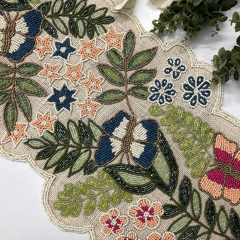 Op de afbeelding: Met kralen versierde tafelloper met geschulpte rand. De loper heeft een beige achtergrond met geborduurde vlinders, bloemen en bladeren in blauw, roze, groen en goud. Het ontwerp is gedetailleerd en ingewikkeld.