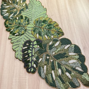 Può includere: Un runner da tavolo decorativo a forma di foglie tropicali decorate con perline. Le foglie sono in varie tonalità di verde, con dettagli bianchi e dorati. Il runner è progettato per assomigliare a foglie di monstera.