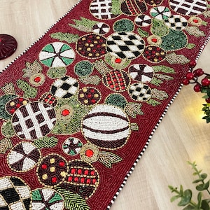 May include: A red beaded table runner with a festive design featuring white, black, green, and gold beads. The design includes a variety of shapes, including circles, squares, and stars. The runner is perfect for adding a touch of holiday cheer to any table.