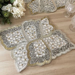 May include: A set of two gold and silver beaded placemats with a geometric design. The placemats are made with small beads and rhinestones.