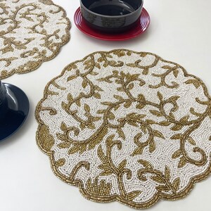 May include: Two white beaded placemats with a gold floral design. The placemats are round and have a scalloped edge.