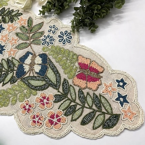 Op de afbeelding: Beaded tafelloper met een geschulpte rand. De loper heeft een bloemen- en bladerontwerp in roze, blauwe, groene en beige tinten. Het ontwerp bevat bloemen, vlinders en stervormige accenten. De loper is gemaakt van een natuurlijk gekleurde stof.
