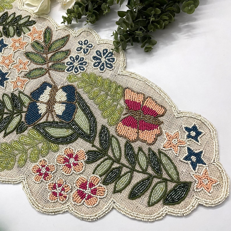 Op de afbeelding: Beaded tafelloper met een geschulpte rand. De loper heeft een bloemen- en bladerontwerp in roze, blauwe, groene en beige tinten. Het ontwerp bevat bloemen, vlinders en stervormige accenten. De loper is gemaakt van een natuurlijk gekleurde stof.