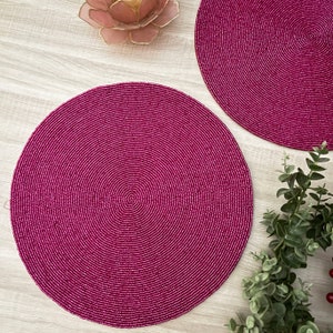May include: Two round pink beaded placemats on a light wood surface. The placemats are made of small pink beads woven together in a circular pattern.