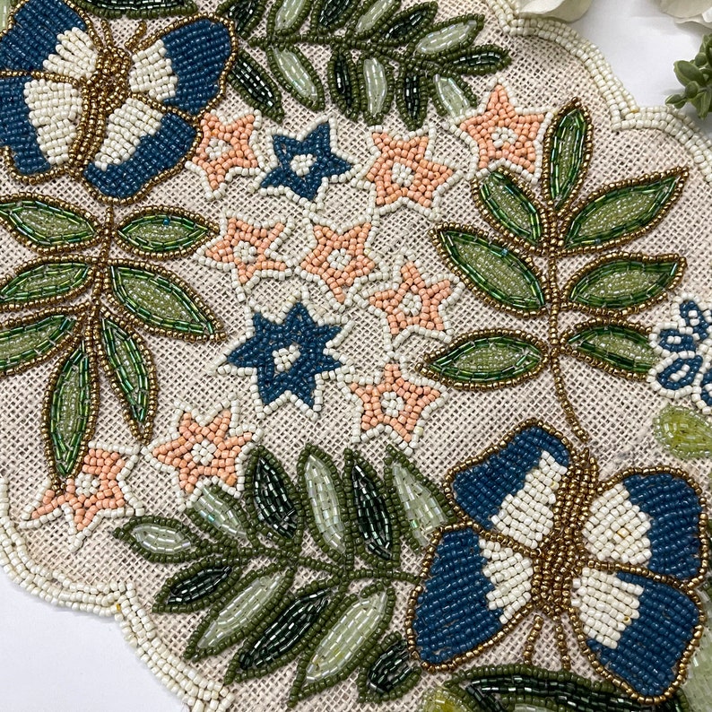Op de afbeelding: Geborduurde tafelloper met een bloemenontwerp. De loper heeft een beige achtergrond met geborduurde bladeren, bloemen en vlinders in tinten blauw, groen en perzik. De randen zijn geschulpt en geborduurd.