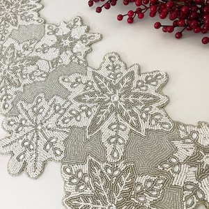 Puede incluir: Un camino de mesa de cuentas blancas y plateadas con un diseño de copo de nieve. El camino de mesa está hecho de pequeñas cuentas redondas que se cosen juntas para crear un patrón delicado e intrincado.