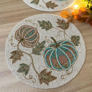 Op de afbeelding: Een ronde, witte placemat met kralen met een ontwerp van twee pompoenen en bladeren in tinten bruin, groen en blauw.