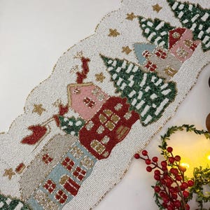 Könnte beinhalten: Weißer Perlen-Tischläufer mit einer Winterdorfszene. Der Läufer zeigt Häuser, Bäume und Sterne in Rot-, Grün-, Rosa- und Goldtönen.