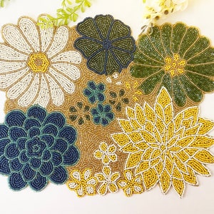 Könnte beinhalten: Eine geperlte Tischunterlage mit Blumenmuster. Die Unterlage zeigt verschiedene Blumen in den Farben Weiß, Gelb, Grün und Blau.