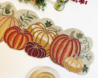 May include: A beaded table runner with a fall theme featuring five pumpkins in shades of orange, yellow, and brown. The pumpkins are surrounded by green leaves and vines.