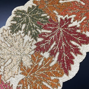 May include: Beaded table runner featuring a design of autumn leaves in shades of red, orange, green, and white.