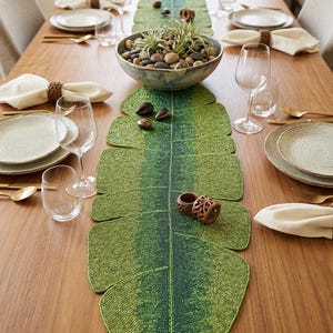 May include: A long, leaf-shaped table runner crafted from small green and yellow beads, featuring a dark green central vein. The runner is placed on a wooden dining table set with white plates, clear glassware, gold cutlery, and cream napkins.