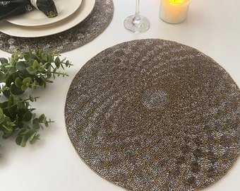 May include: A round beaded placemat with a gold and silver pattern. The placemat is made of small beads that are woven together to create a intricate design.