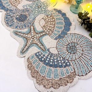 Pode incluir: Um caminho de mesa com contas com um design costeiro, com motivos de estrelas do mar e conchas. O caminho de mesa é principalmente branco, azul e castanho, com detalhes de pérolas. Mede cerca de 30 cm de largura e 152 cm de comprimento.