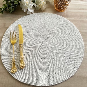 Op de afbeelding: Ronde placemat van witte kralen met een gouden vork en mes erop.