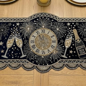 Puede incluir: Camino de mesa azul marino con adornos de cuentas doradas y plateadas. Presenta un reloj, copas de champán, fuegos artificiales y las palabras "Cheers!" y "Happy New Year". Borlas decorativas cuelgan de los extremos. Un adorno festivo.