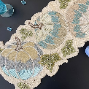 Könnte beinhalten: Ein geperlter Tischläufer mit einem Design aus drei Kürbissen und Blättern. Die Kürbisse sind beige, blau und grün. Die Blätter sind grün.