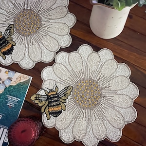Op de afbeelding: Twee witte parel placemats met een madeliefje ontwerp en een bij in het midden. De placemats liggen op een houten tafel.