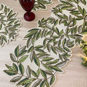 Puede incluir: Camino de mesa y manteles individuales con diseño de hojas. Las hojas son verdes, grises y marrones, sobre un fondo blanquecino. Se ve una copa de vino burdeos. El conjunto está sobre una superficie de madera clara.