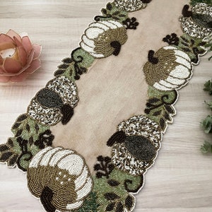 May include: A beige table runner with a beaded design featuring pumpkins, leaves, and vines. The beads are in shades of brown, white, and green.