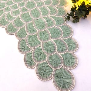 May include: A beaded table runner in a repeating teardrop pattern. The runner features a light green color with a gray border and white accents. The runner is made of small beads and is suitable for home decor.