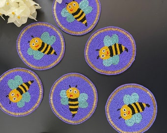 May include: Six purple and gold beaded coasters with a bee design. Each coaster features a yellow bee with black stripes and a blue and green body. The bees have smiling faces.