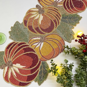 May include: A beaded table runner with a autumnal theme featuring pumpkins and leaves in shades of orange, red, green, and gold.
