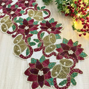 Può includere: Una tovaglia natalizia con perline con stelle di Natale, campane e nastri. La tovaglia ha uno sfondo bianco con accenti bordeaux, verdi e dorati. Le stelle di Natale sono di un colore bordeaux intenso e le campane sono dorate.
